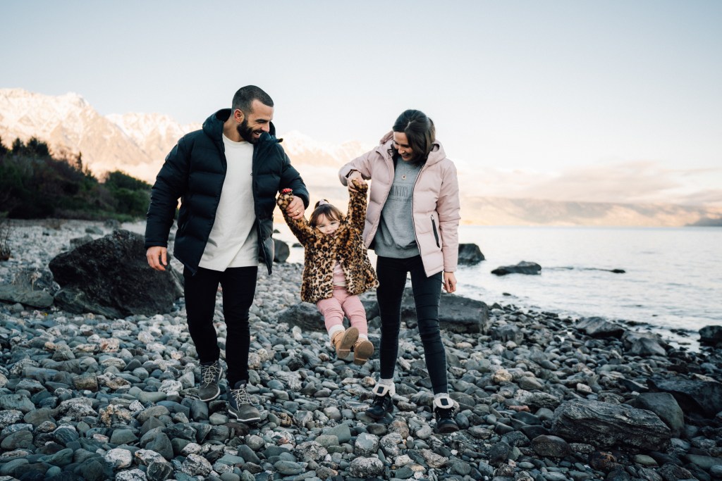 A travel photographer captures an image of a male and a female are walking around the edge of a lake in Queenstown holding the hands of a small child. 